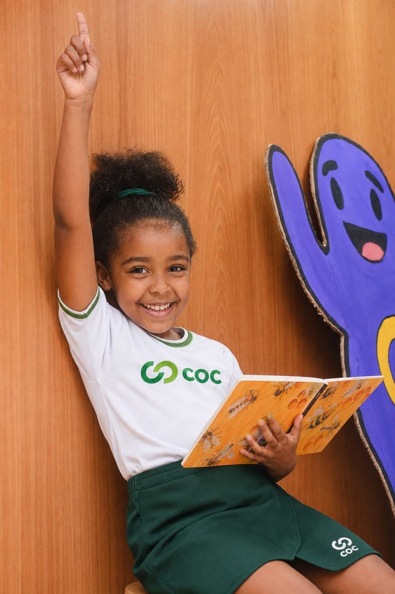 Aluna da Educação Infantil do COC com um livro, de mão levantada