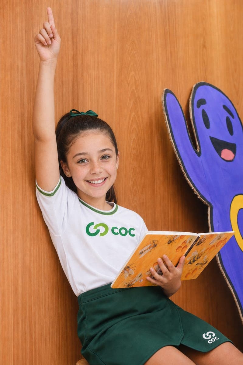 Aluna da Educação Infantil do COC com um livro aberto, de mão levantada